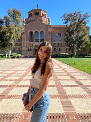Photo of Claire smiling at the camera outside of Powell Library wearing jeans and a white shirt on a sunny day. 