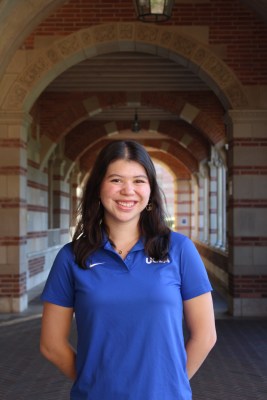 Kathryn wears a blue UCLA polo shirt and smiles at the camera while standing outside of Royce Hall. 