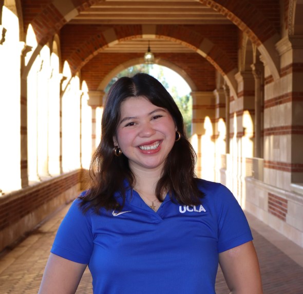 Professional photo of Kathryn smiling at the camera wearing a blue UCLA polo shirt and standing in the hallway outside of Royce Hall.