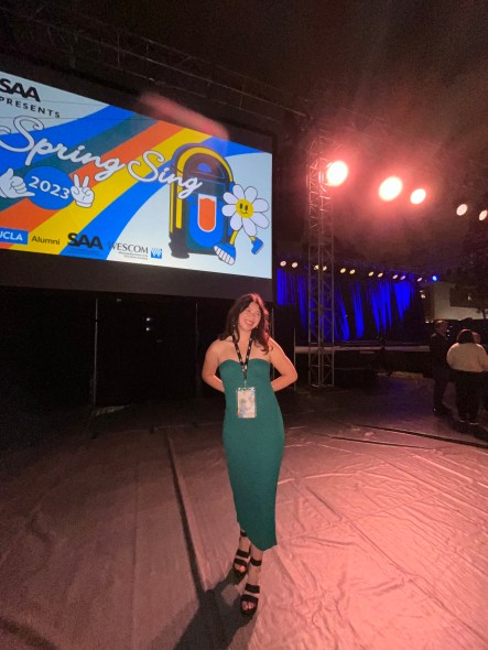 Photo of Kathryn wearing a green dress and event lanyard and standing in front of the UCLA Spring Sing 2023 event sign.