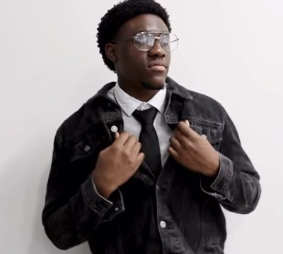 Divine wearing glasses, a tie and denim jacket.