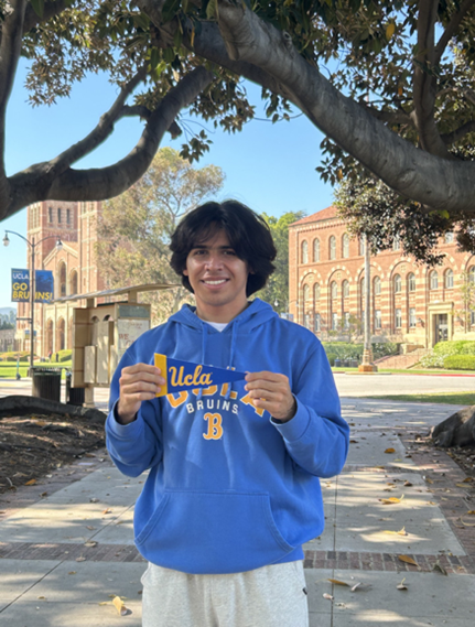 Fabrizio on campus wearing a blue UCLA sweatshirt and holding a UCLA flag with Royce Hall in the background.