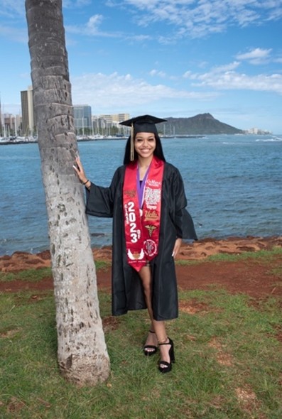 Nay wearing a cap and gown and high school graduation regalia with the bay in the background.  