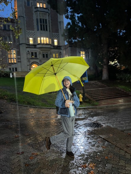 Namuun on a rainy evening on campus holding a yellow umbrella and posing in front of Kerckhoff Hall at UCLA. 